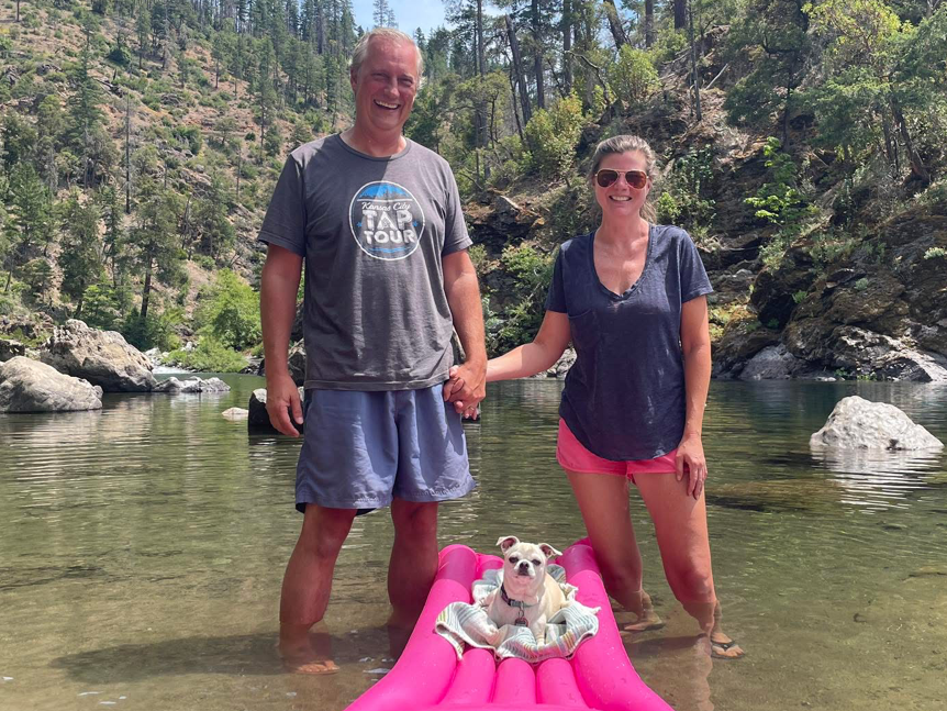 A happy couple standing in shallow water, holding hands, with a small dog on a pink float nearby. The background features a rocky terrain and lush greenery.