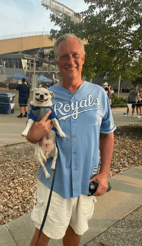 A smiling man wearing a light blue Royals jersey holds a small dog in a blue outfit, with a sports venue in the background.